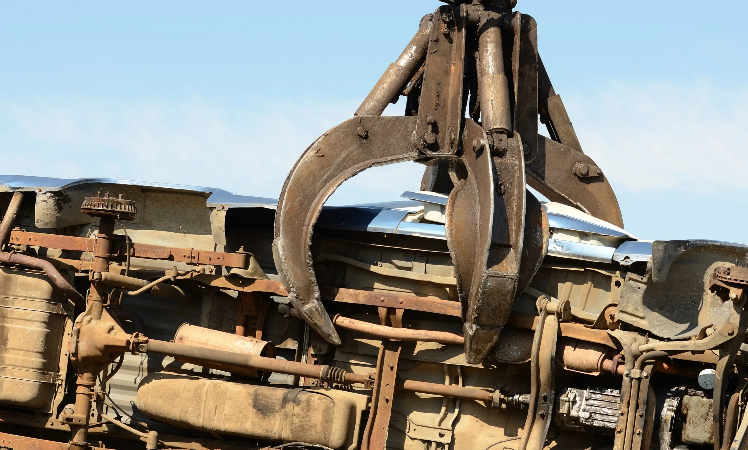 Heavy industrial crane claw lifting and crushing a scrap car in a recycling yard.